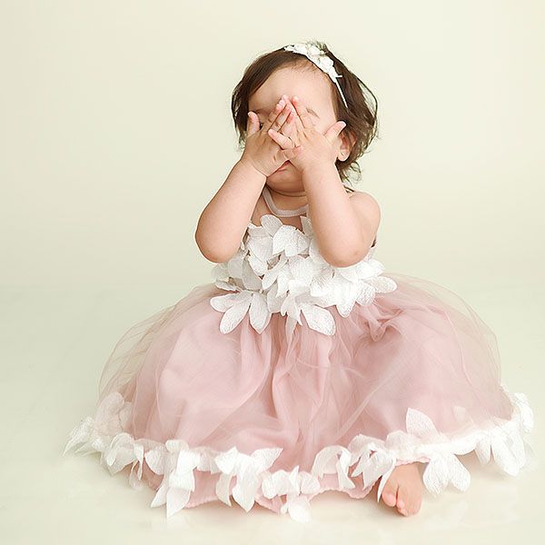 A small child in a pink tulle dress with white bows covers their face while sitting in a whimsical pose.