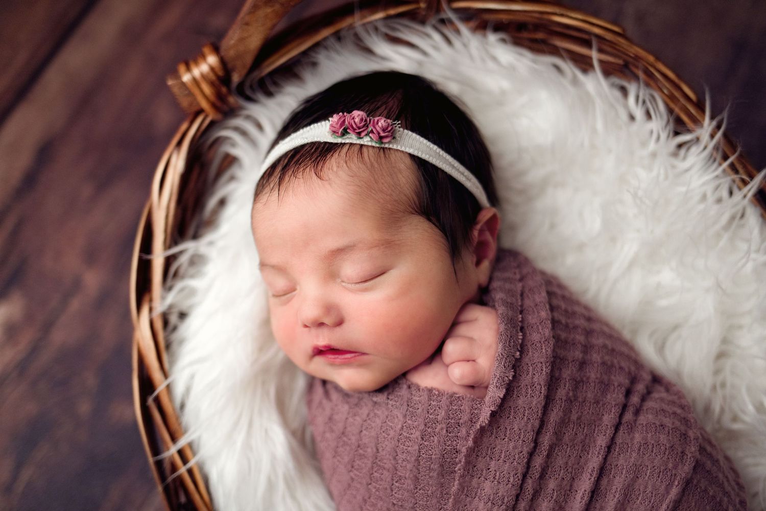 Newborn baby sleeping peacefully on soft white fur in brown wrap with headband during professional photoshoot.