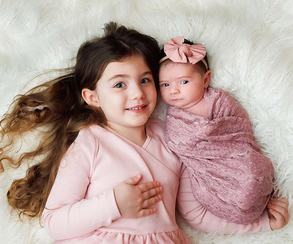 Two children in pink outfits lying on white fluffy rug in coordinated sibling photo session.