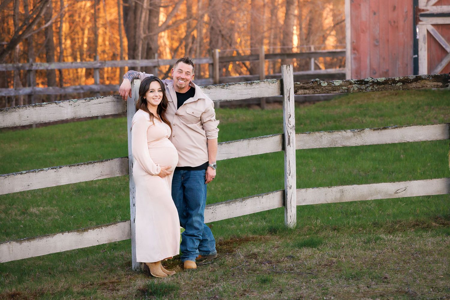 Pregnant couple posing by white fence at golden hour with red barn backdrop during maternity photo session.