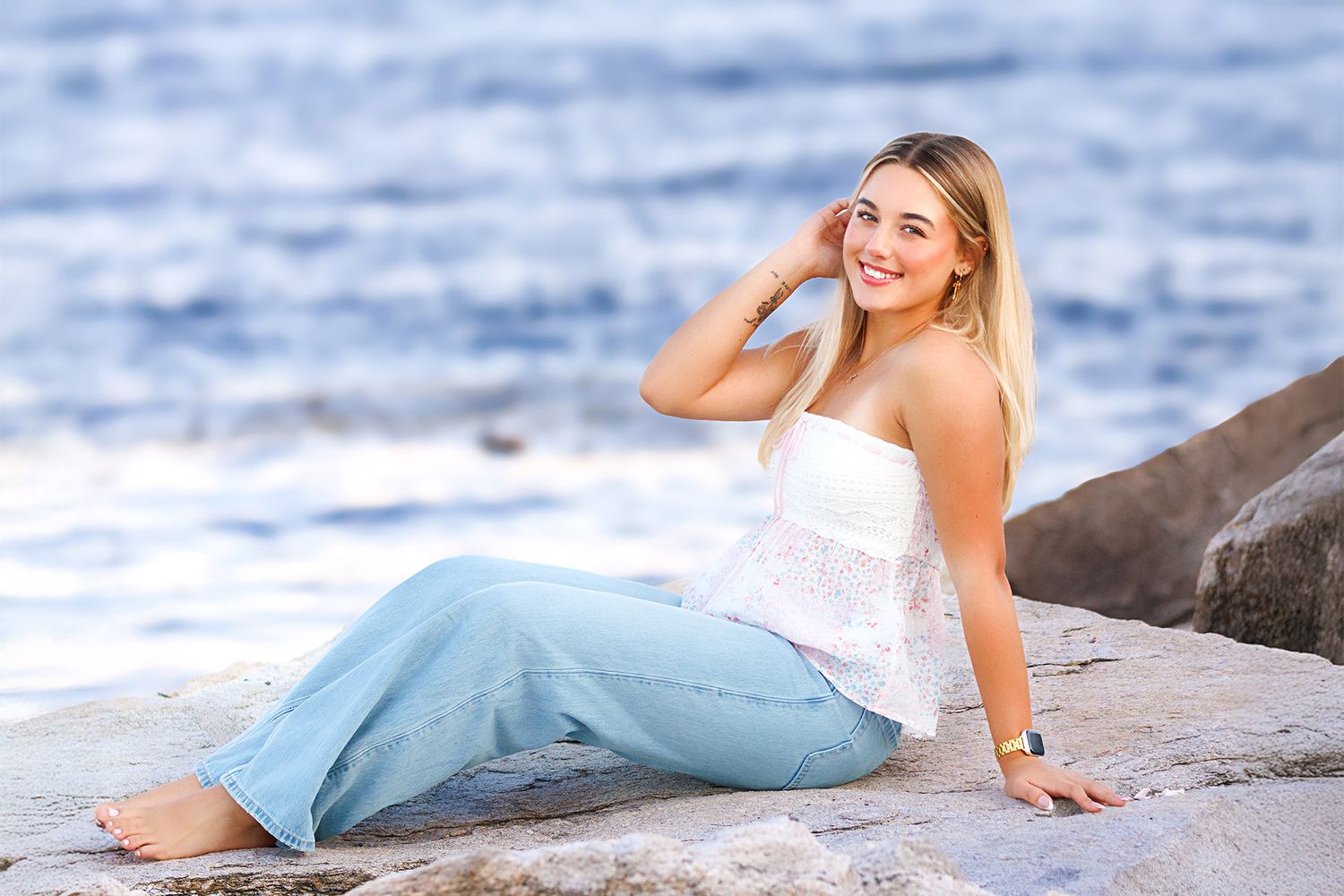 Young blonde woman in white top and light blue pants sitting on rocks by the ocean, smiling at camera.