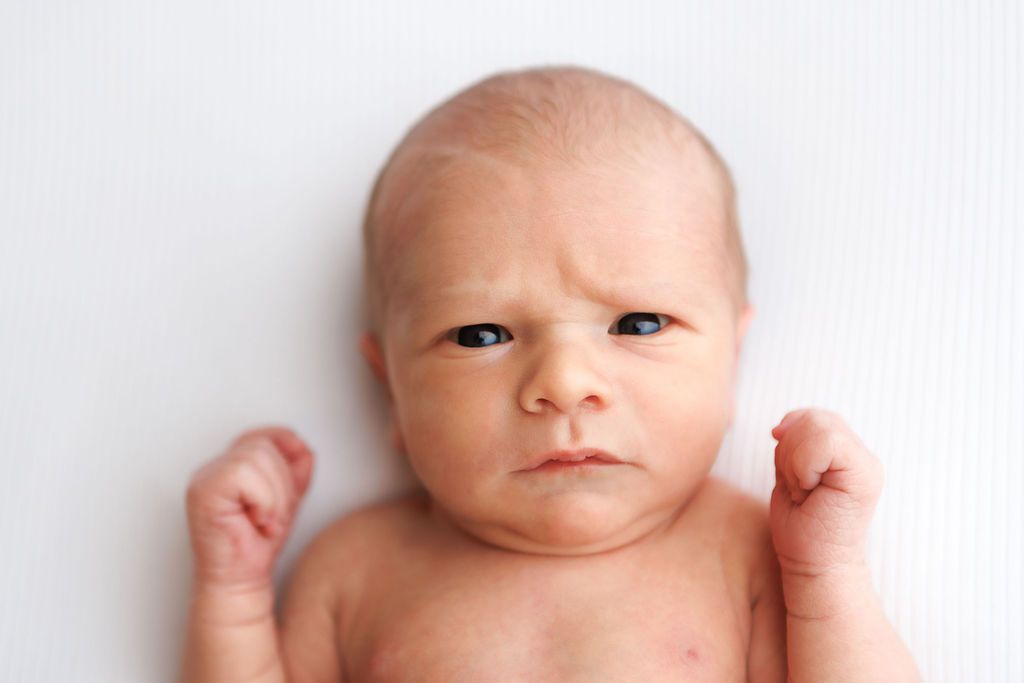 Newborn baby with furrowed brow and raised fists lying on white background, making an adorably grumpy expression.