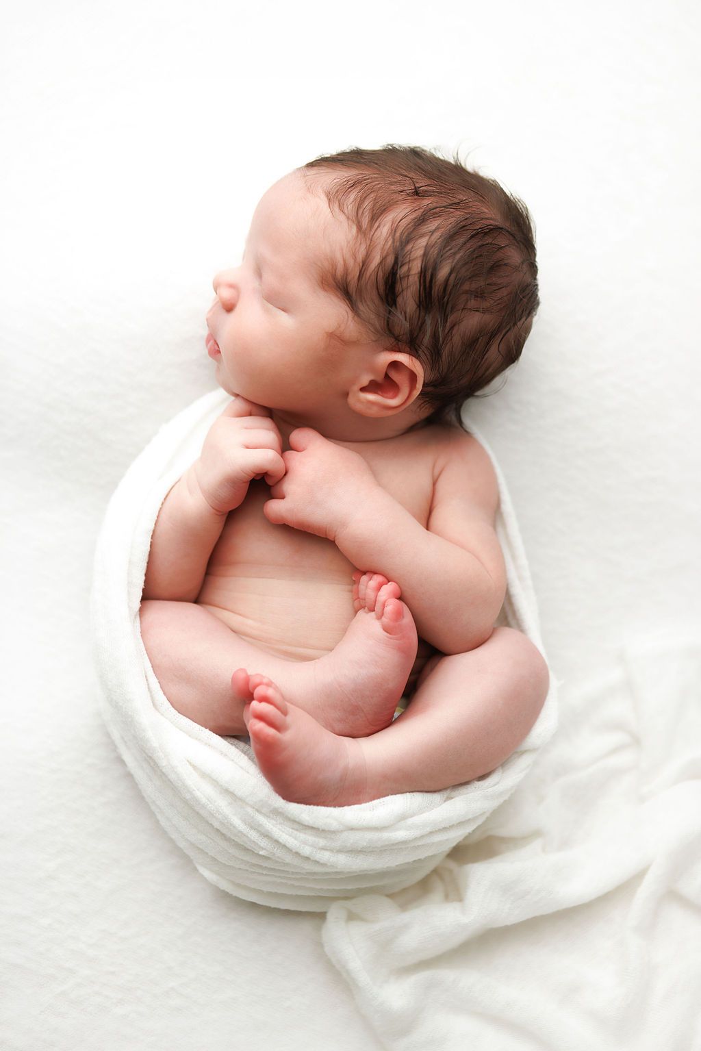 Peaceful newborn baby sleeping curled up in a white swaddle wrap on a soft white blanket.