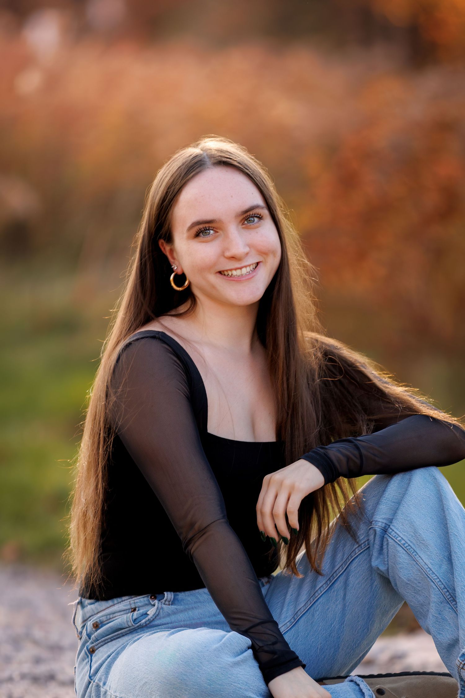 Young woman with long brown hair smiling outdoors, wearing black top and jeans, seated against autumn foliage background.