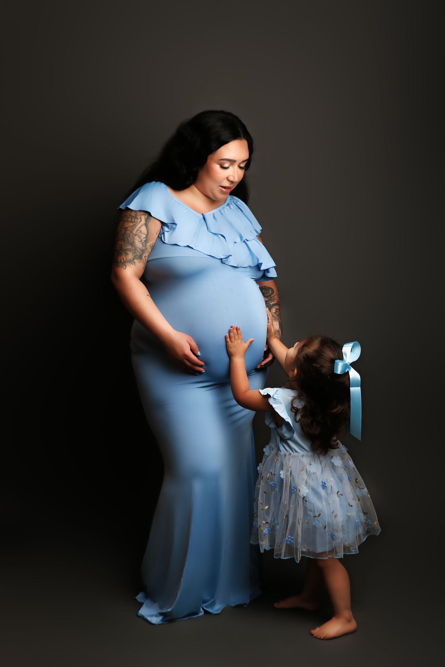 A touching moment captured in a dark studio shows a mother in a light blue dress kneeling beside her child in a tulle dress.