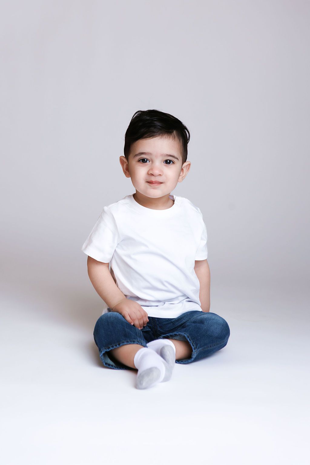 Cute toddler boy sitting cross-legged on white background, wearing white t-shirt, jeans, and white socks.