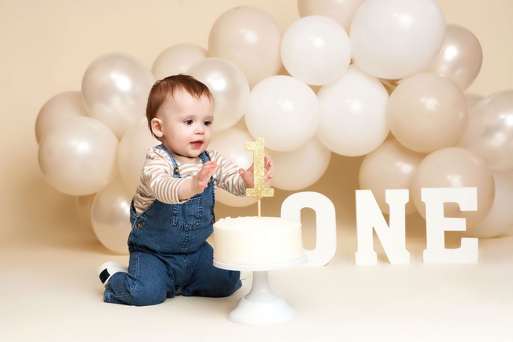 Baby in denim overalls celebrating first birthday with white cake and ONE sign surrounded by neutral balloons.