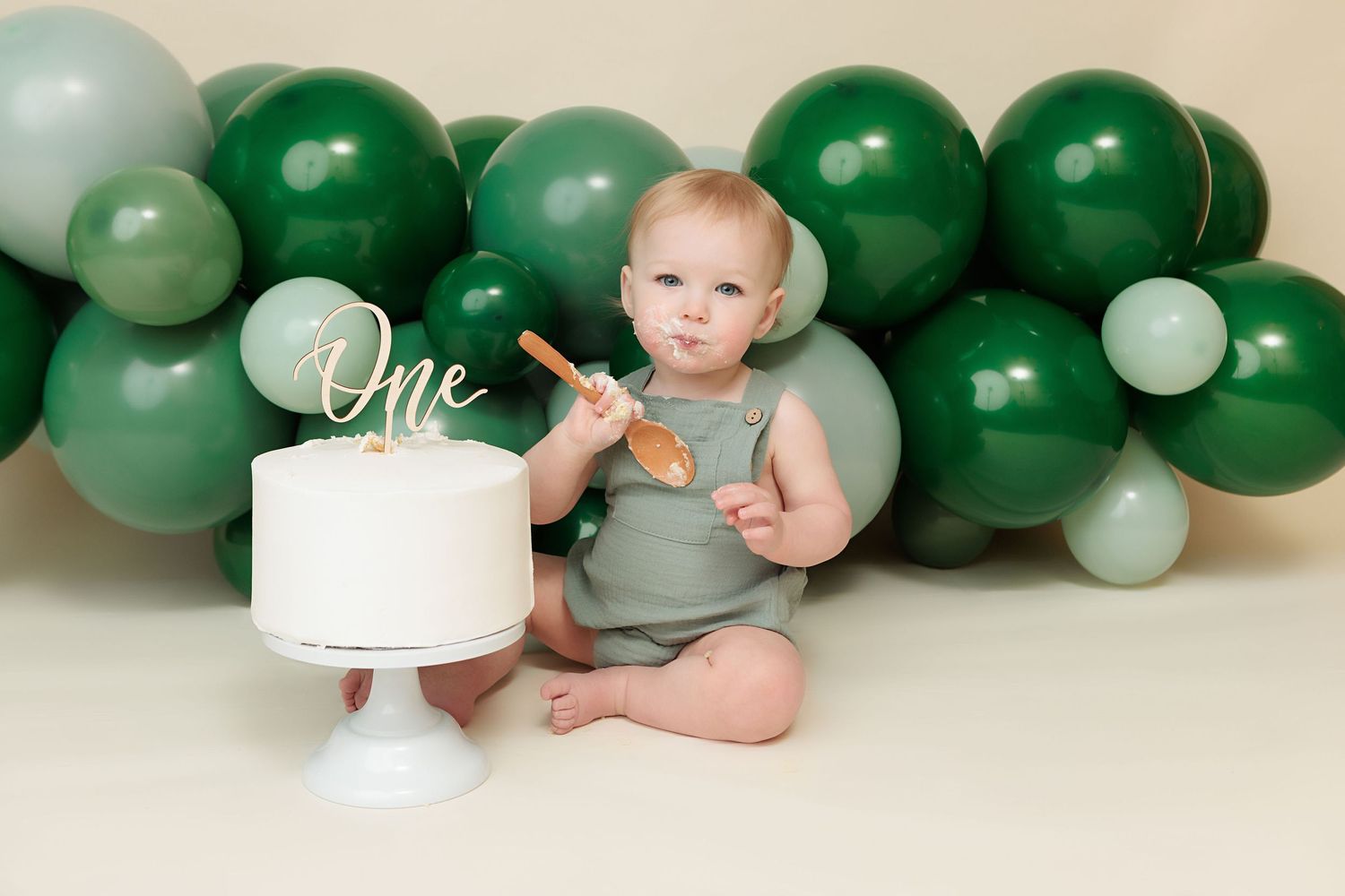 Baby in green romper sits with white cake and wooden spoon, surrounded by green balloon garland for first birthday.