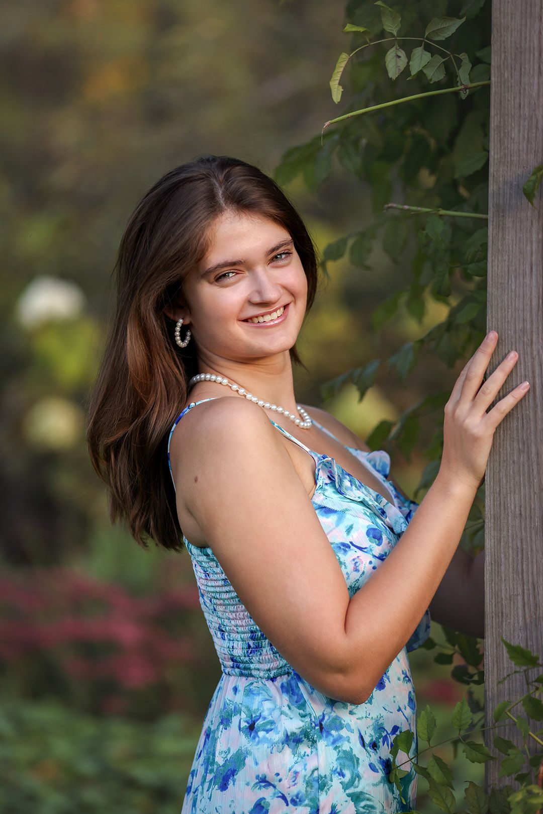 Young woman in blue floral dress and pearl necklace smiling outdoors near wooden post with greenery.