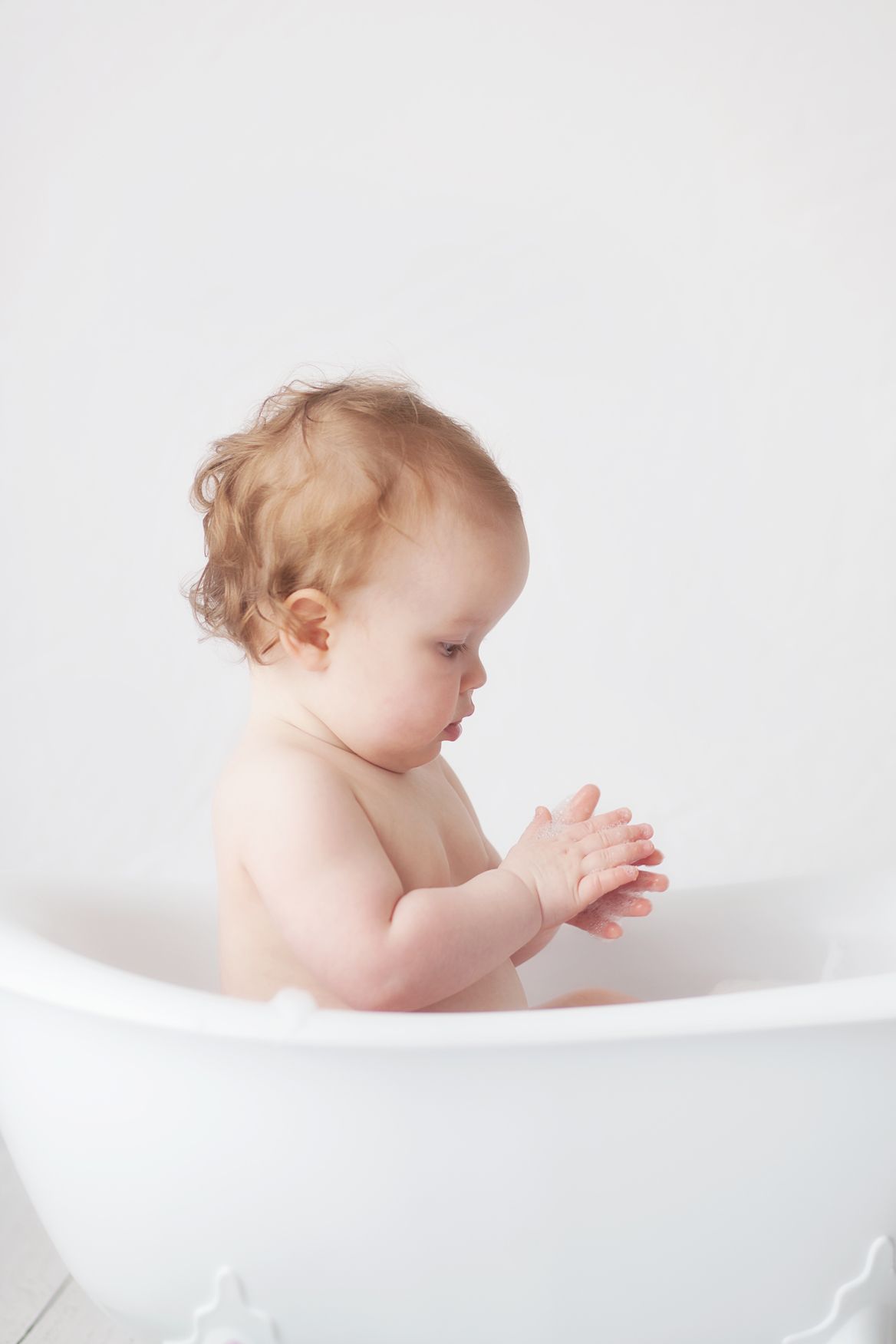 A baby sits in a white bathtub against a light background in a minimalist artistic composition.