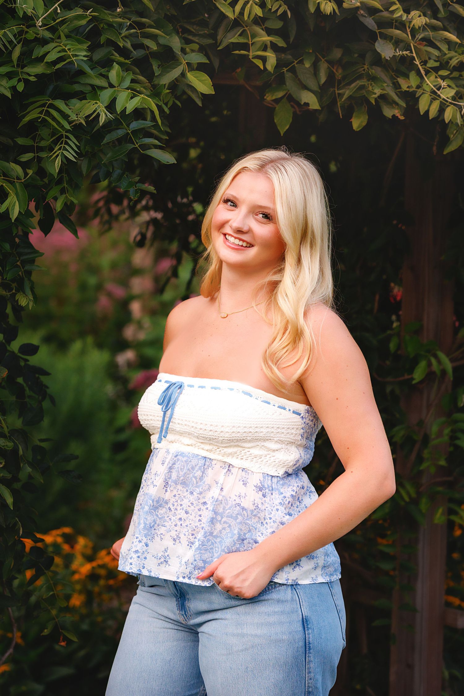 Windham NH Senior in a white lace top and jeans poses outdoors against green foliage in a garden setting.