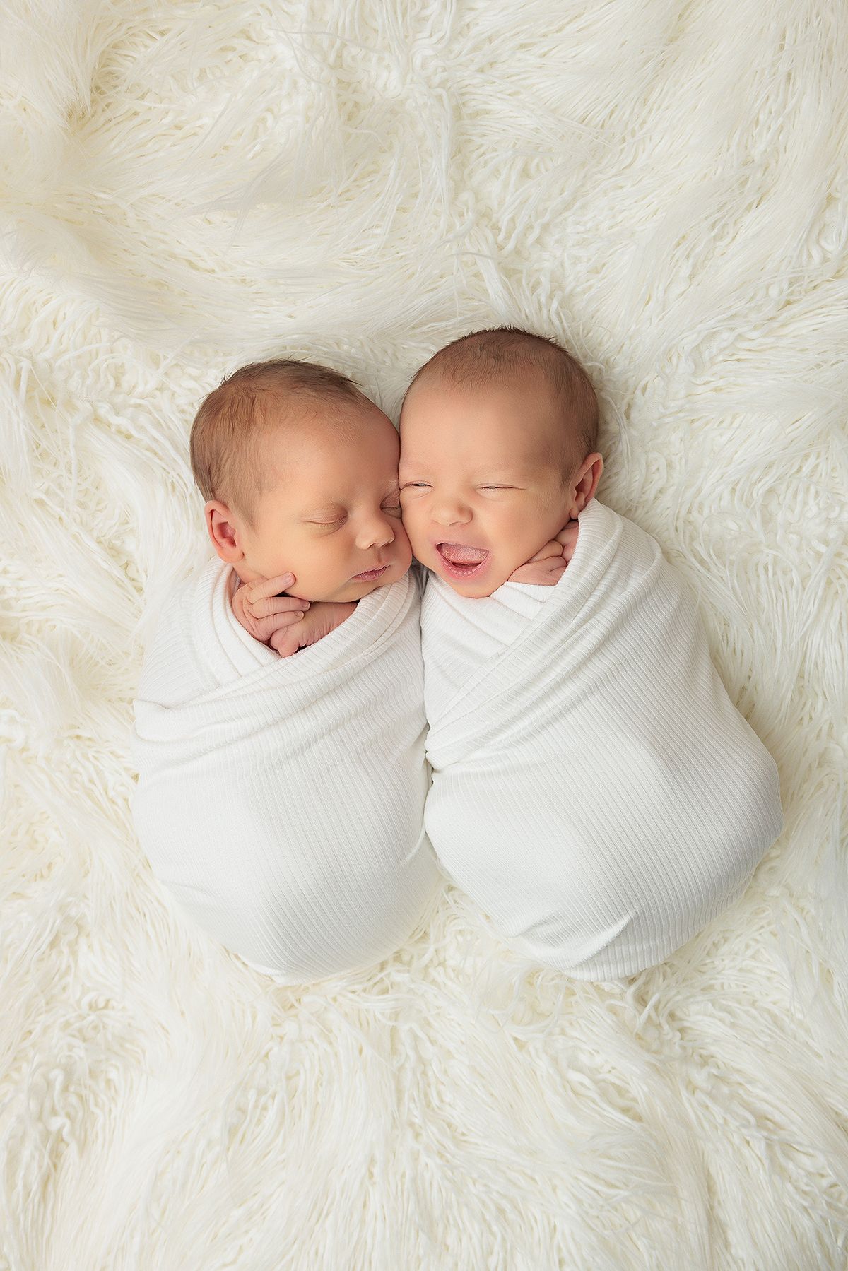 Two sleeping newborn babies swaddled in white blankets lying together on soft white fur.