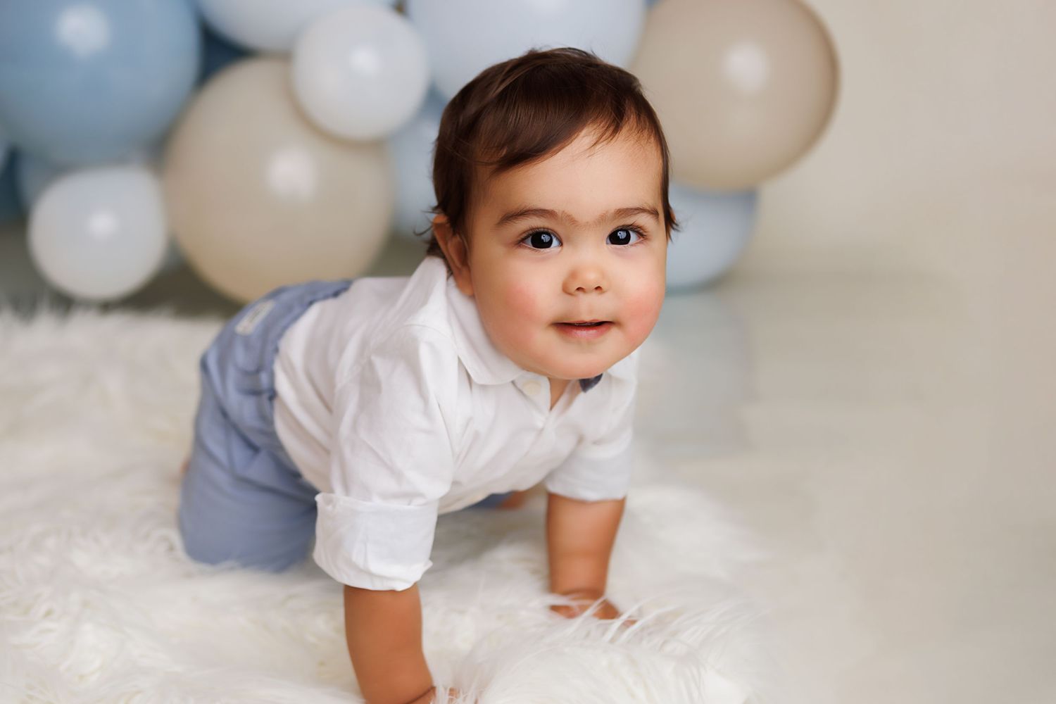 Baby in white outfit crawls and smiles on light gray backdrop with pastel balloons in the background.