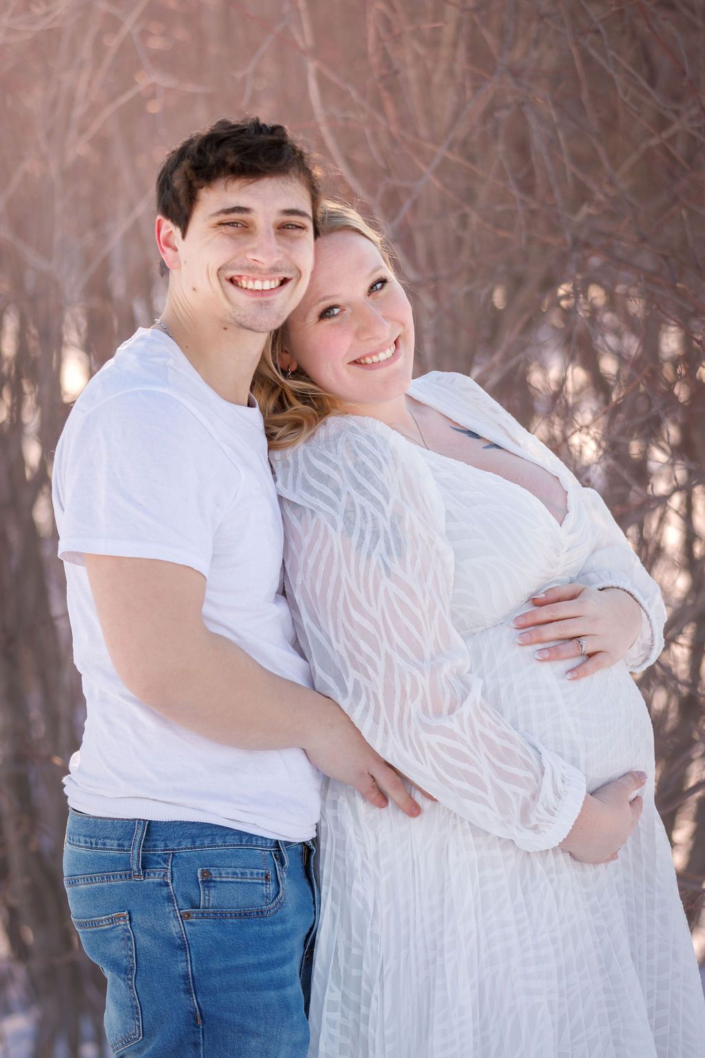 Smiling couple in white outfits posing outdoors in winter, woman in lace maternity gown cradling baby bump.