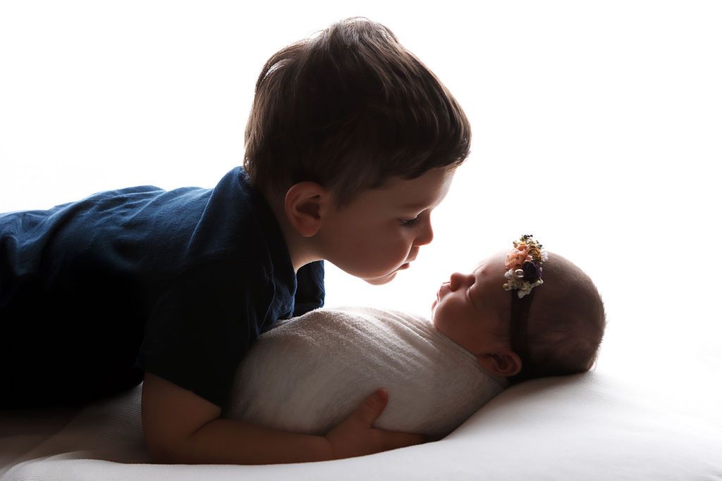 A young boy gently kisses his newborn sibling wrapped in white, captured in a tender siblings portrait.