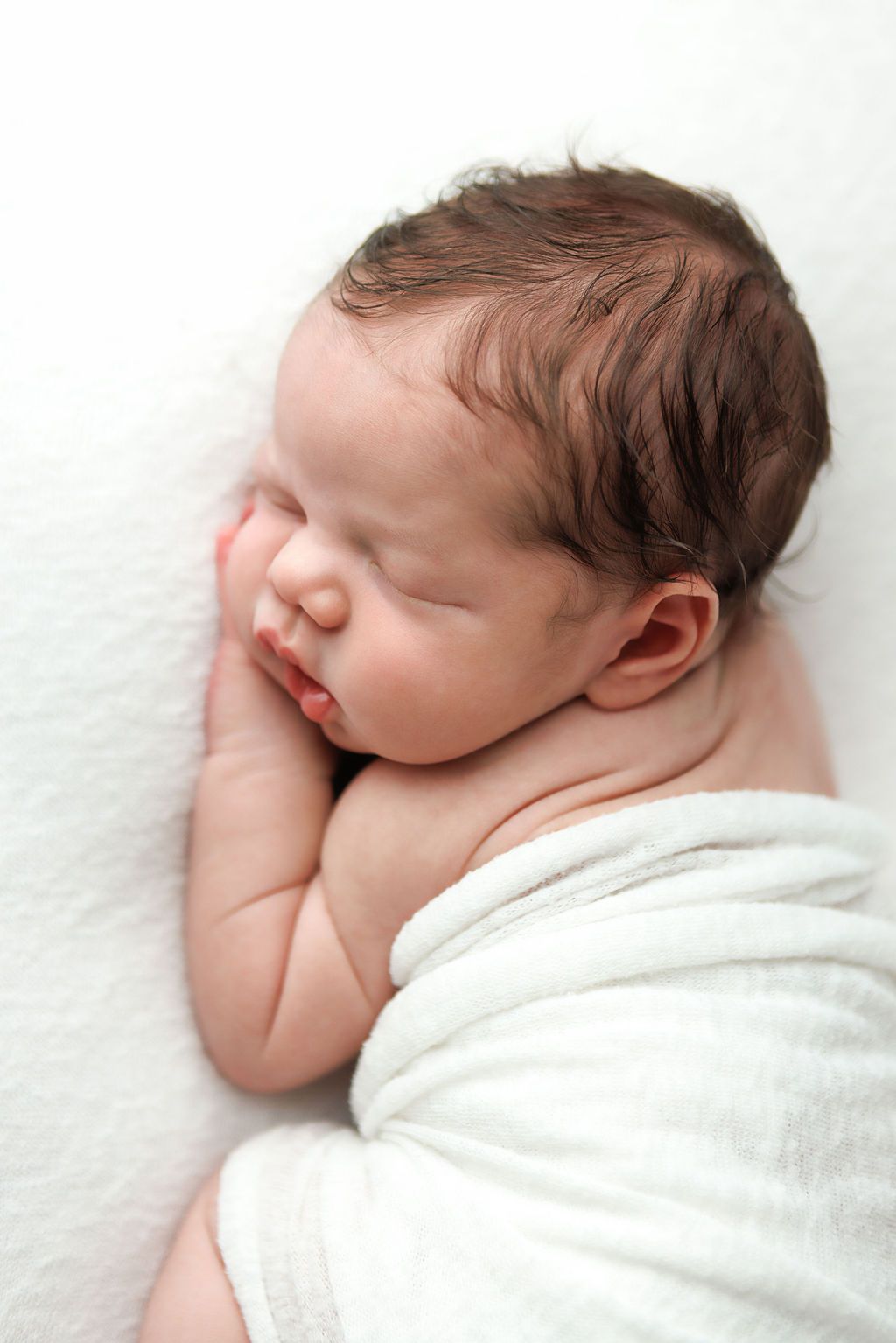 Peaceful newborn baby with dark hair sleeping wrapped in a white swaddle blanket on a soft white background.