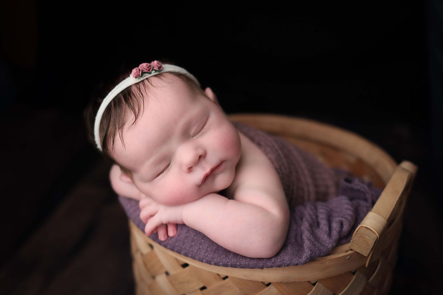 Newborn baby sleeping peacefully in wooden bowl with purple blanket in professional studio setting.
