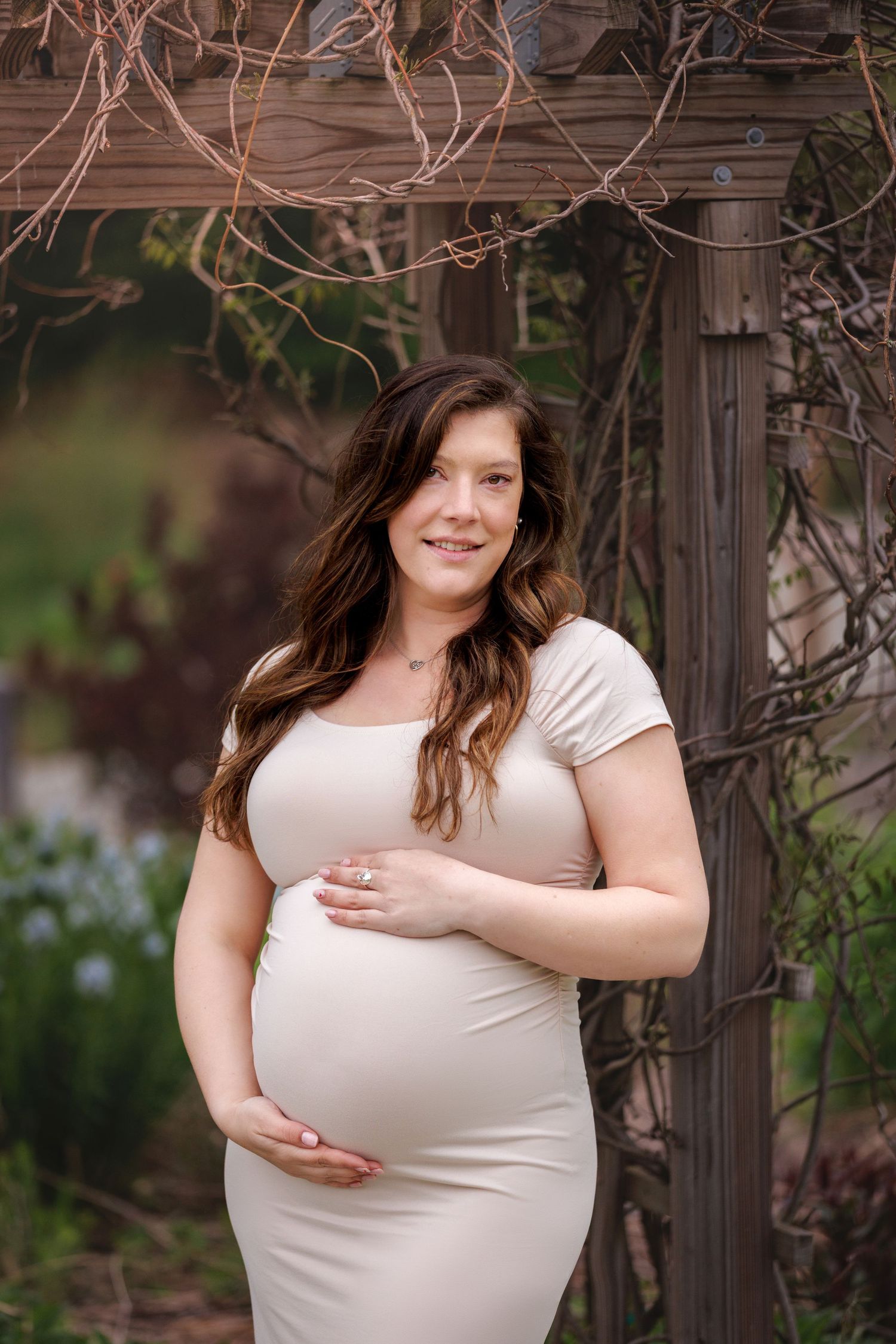 A pregnant woman in a beige dress poses outdoors near a wooden structure with vines and blue flowers in the background.
