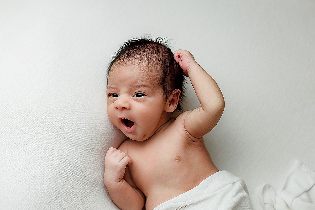 A newborn baby with dark hair lies on white fabric while making an expressive yawning face.