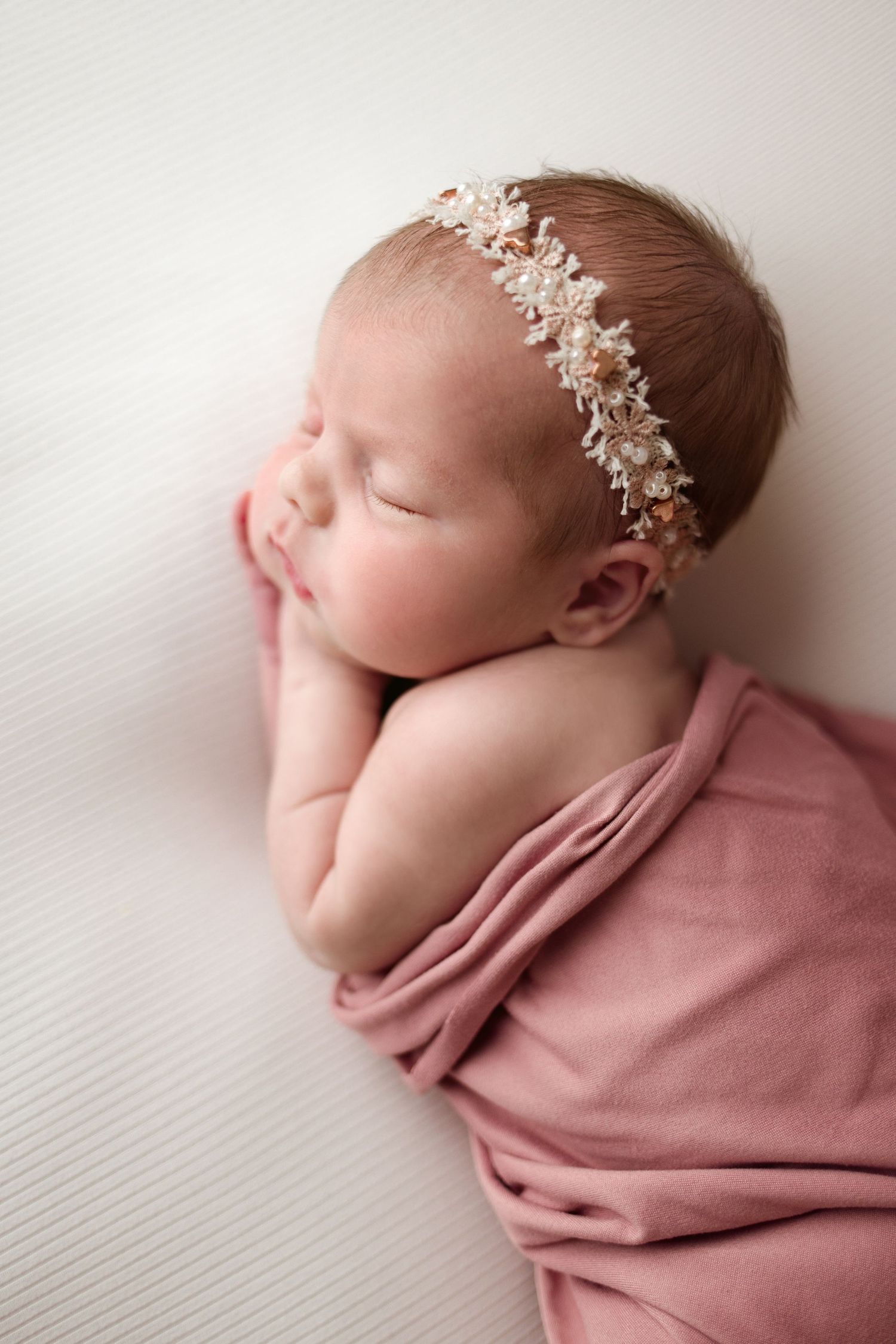 Sleeping newborn wrapped in soft pink fabric wearing delicate floral lace headband during professional photo session.