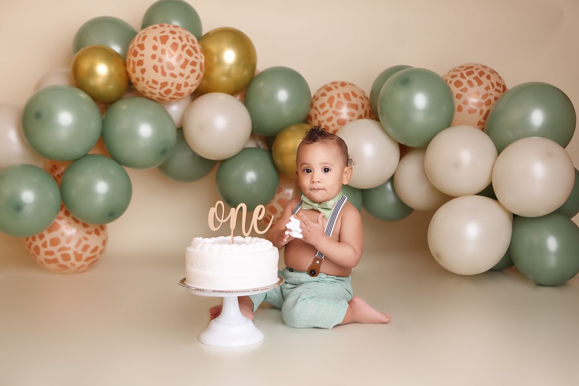 First birthday celebration setup with sage green and gold balloon arch and white cake on pedestal against neutral backdrop.