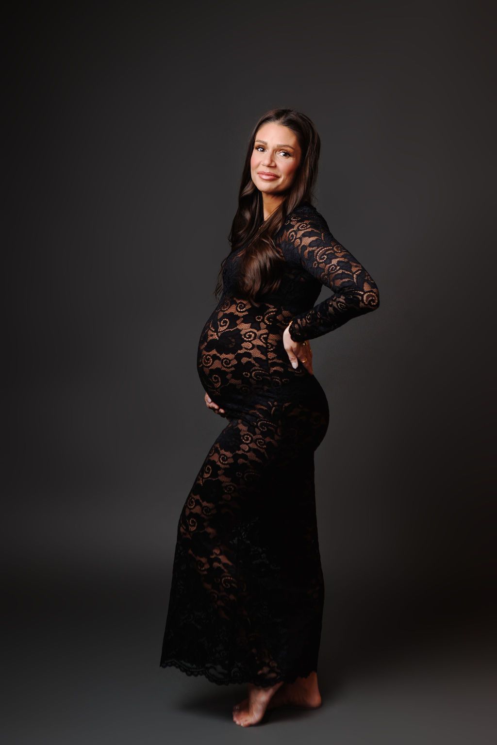 Pregnant woman in elegant black lace gown posing gracefully in a studio, showcasing her baby bump.