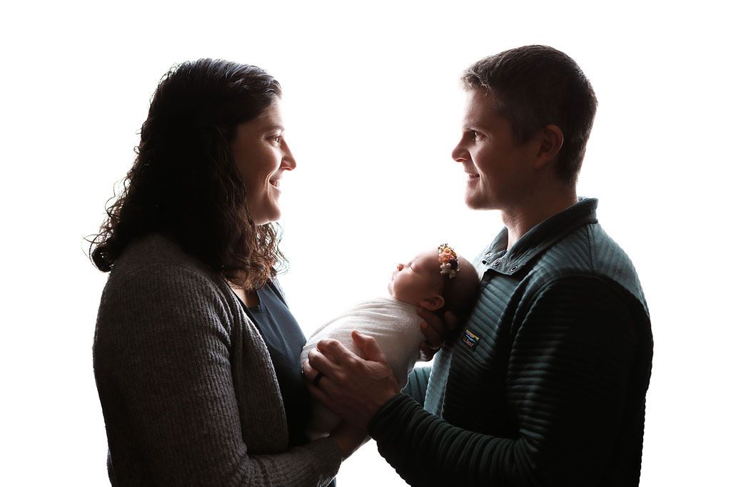 Silhouette of parents holding their swaddled newborn baby against bright white background in tender family portrait.