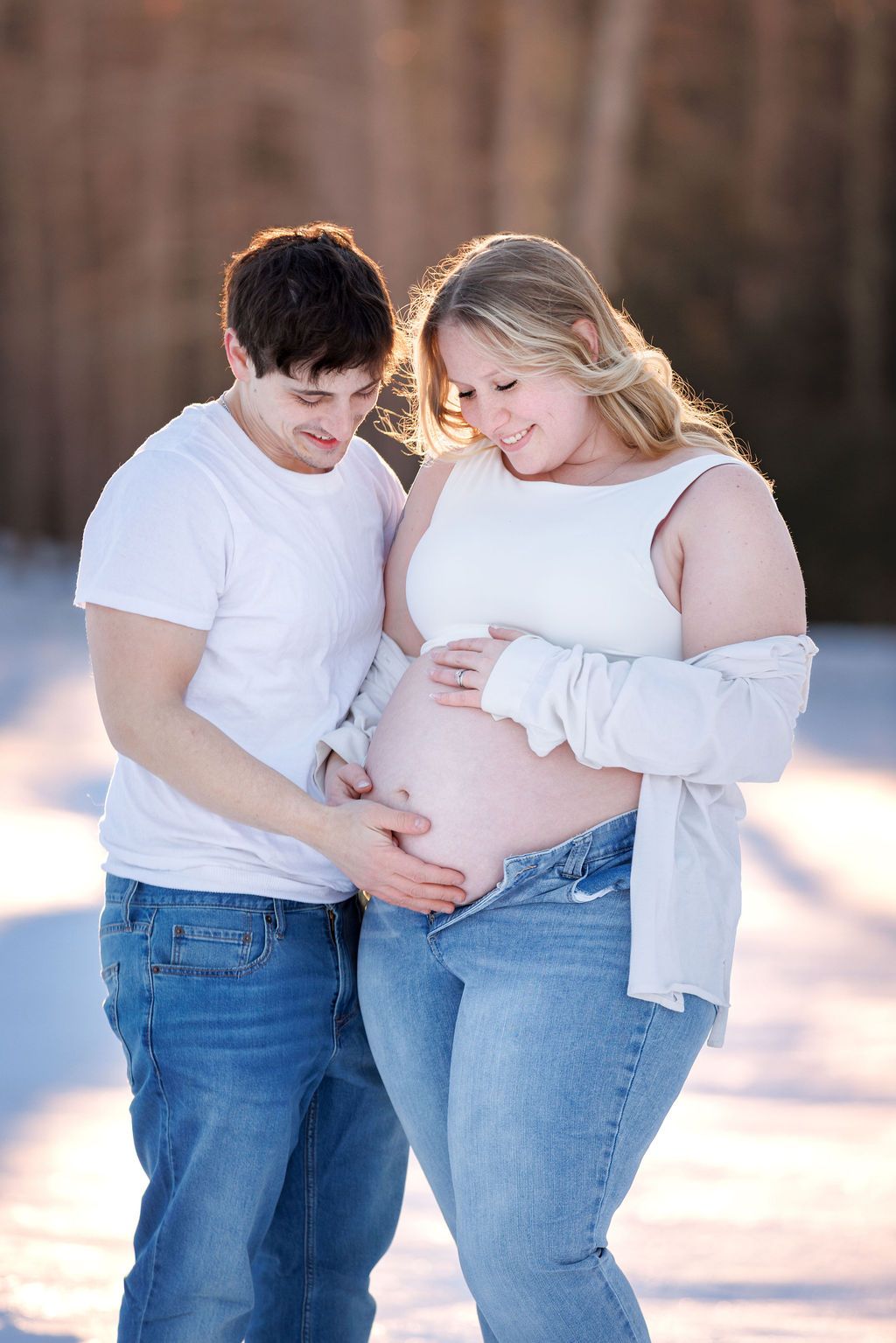 Couple in white tops and jeans cradle pregnant belly outdoors in snowy winter setting with warm golden sunlight.