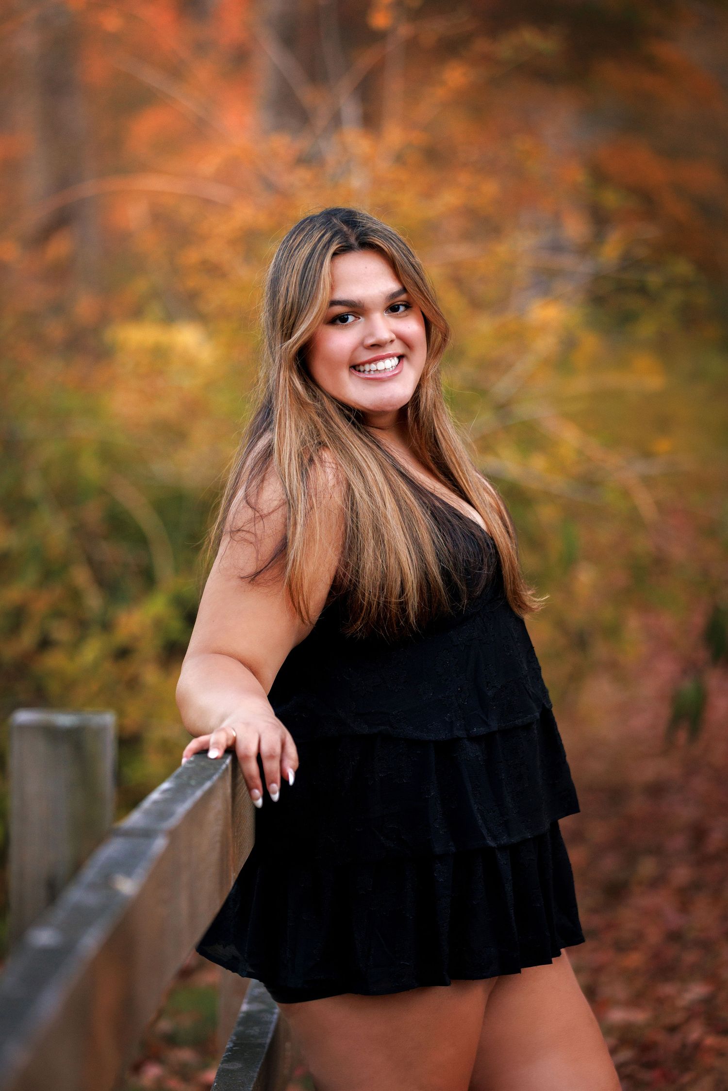 Young woman in black dress smiling outdoors by wooden fence with colorful autumn foliage background.