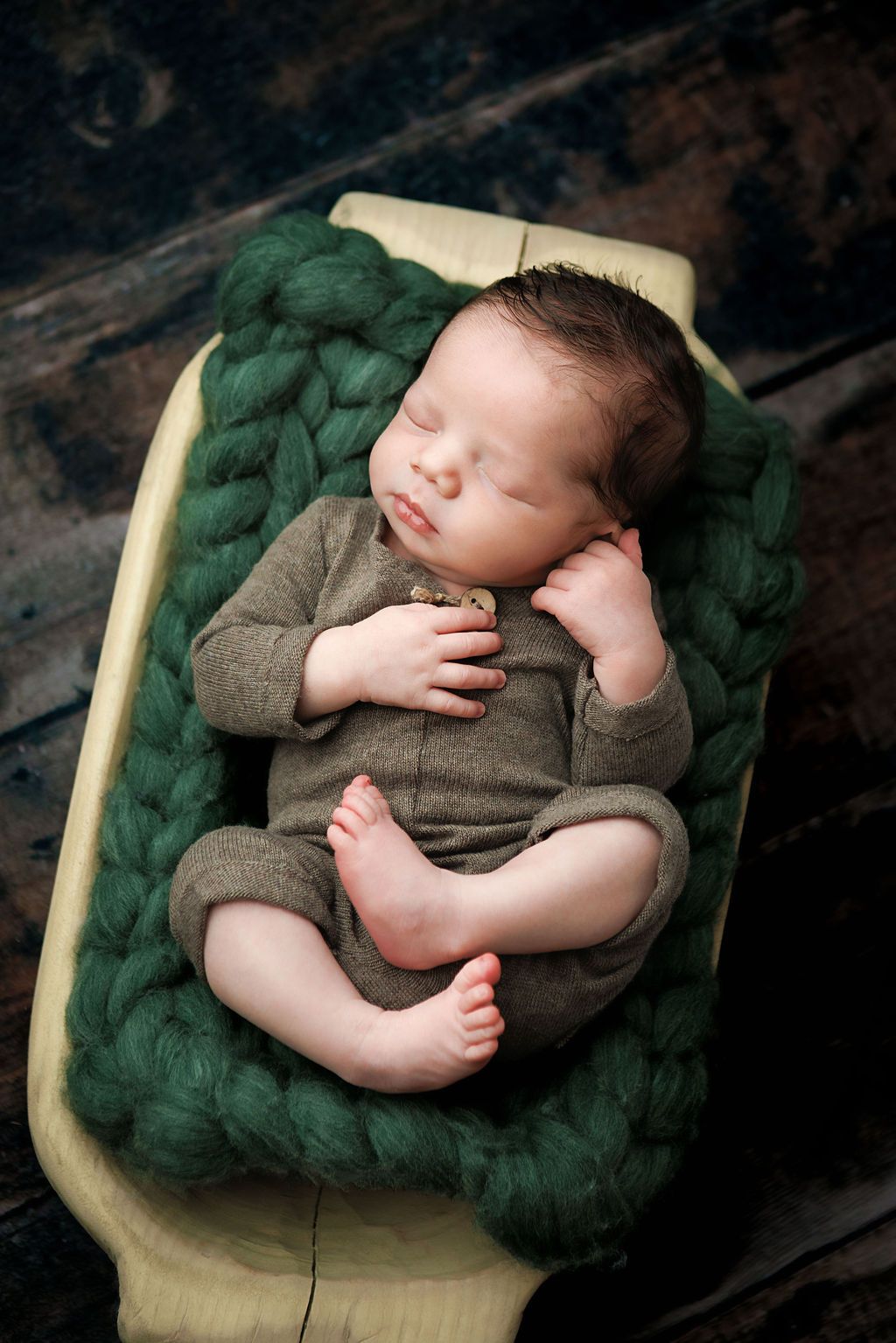 Sleeping newborn baby in olive romper resting on green chunky knit blanket inside a small wooden bed.
