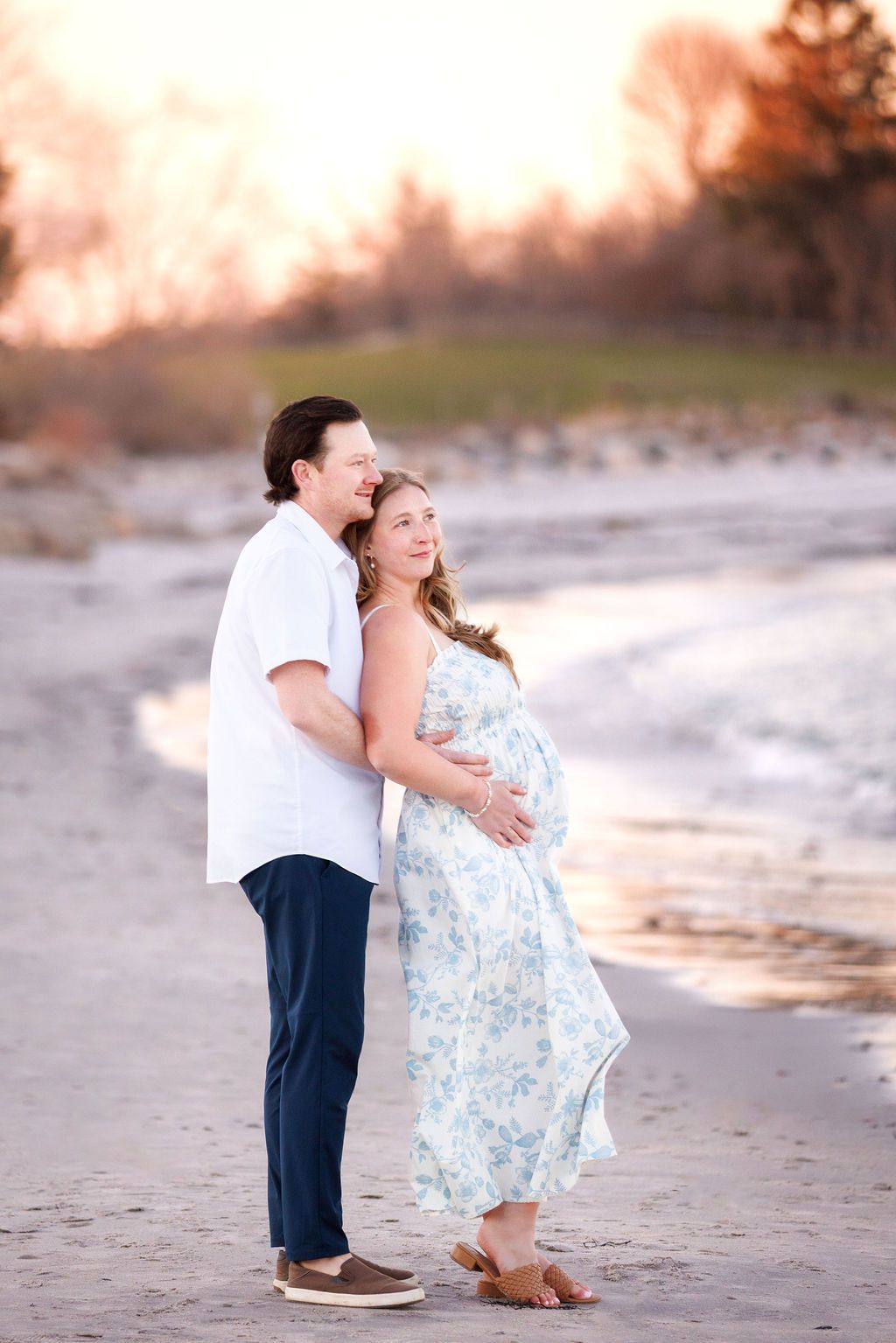 Pregnant couple embracing on a sandy beach at golden hour, woman in floral dress cradling baby bump.