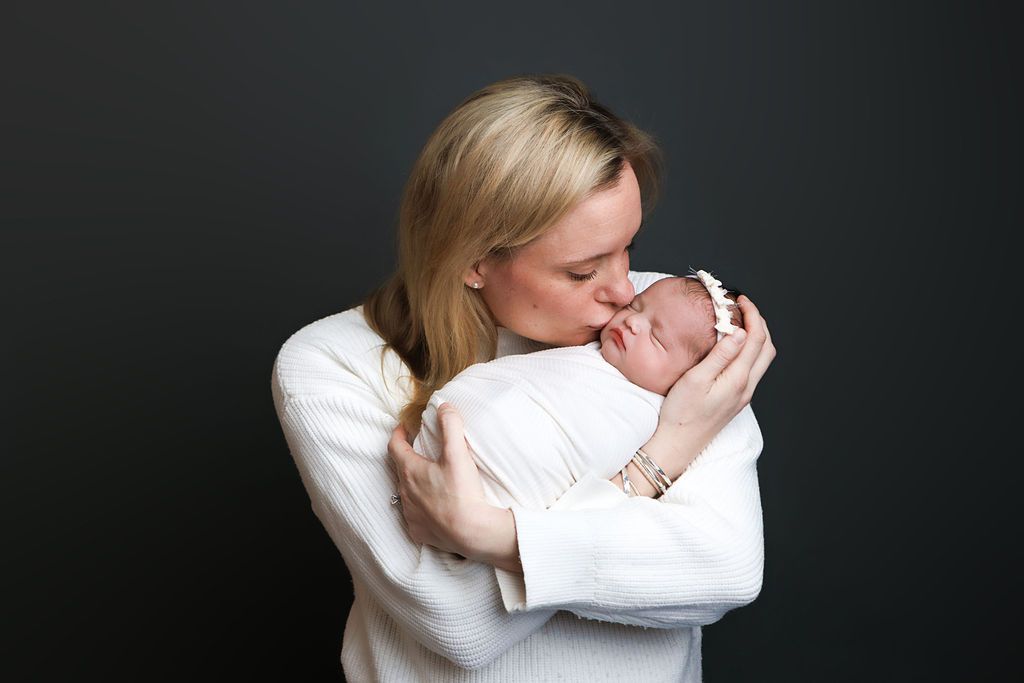 A blonde mother tenderly kisses her sleeping newborn baby wrapped in white against a dark background.