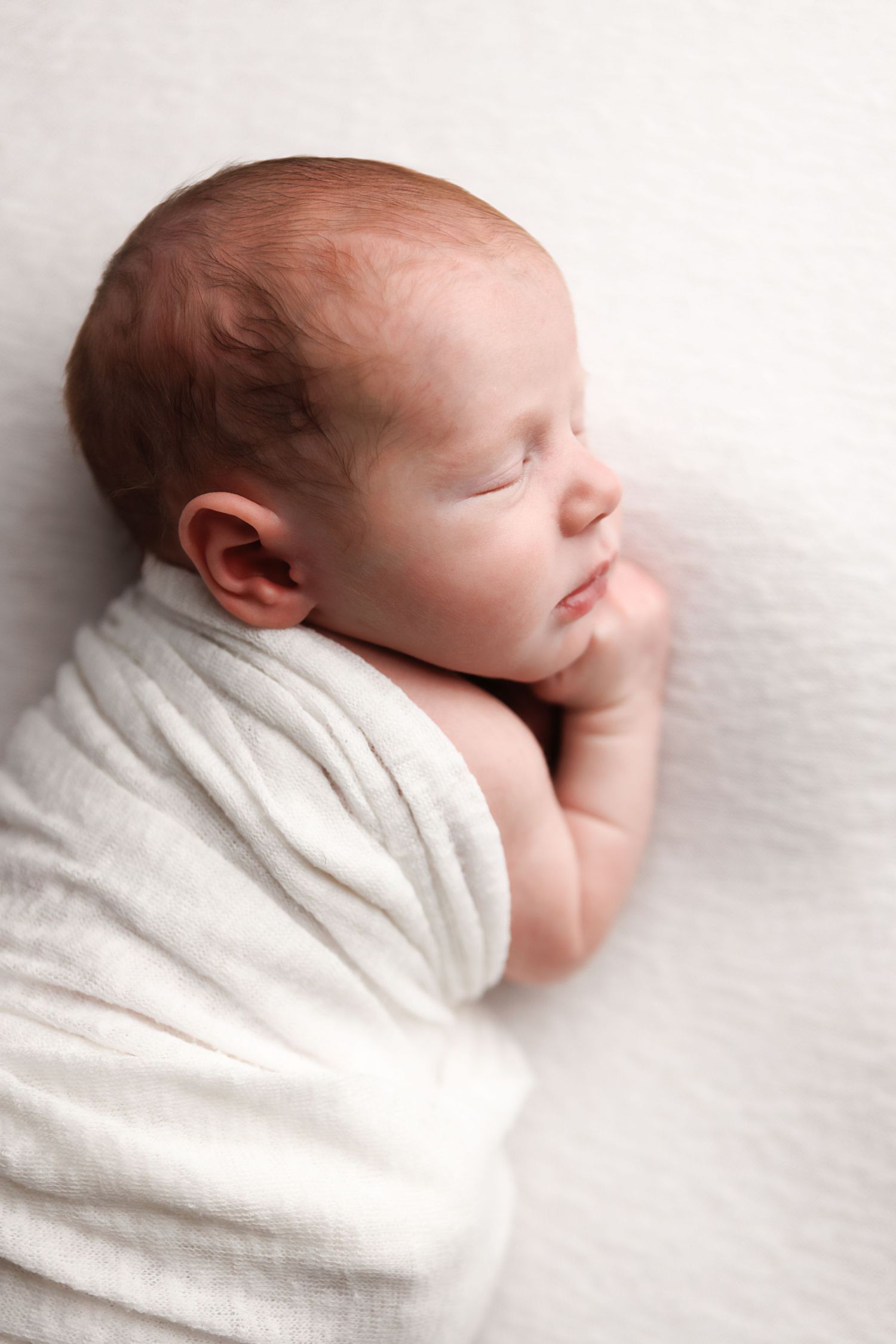 A sleeping newborn baby swaddled in white cloth against a soft white background.