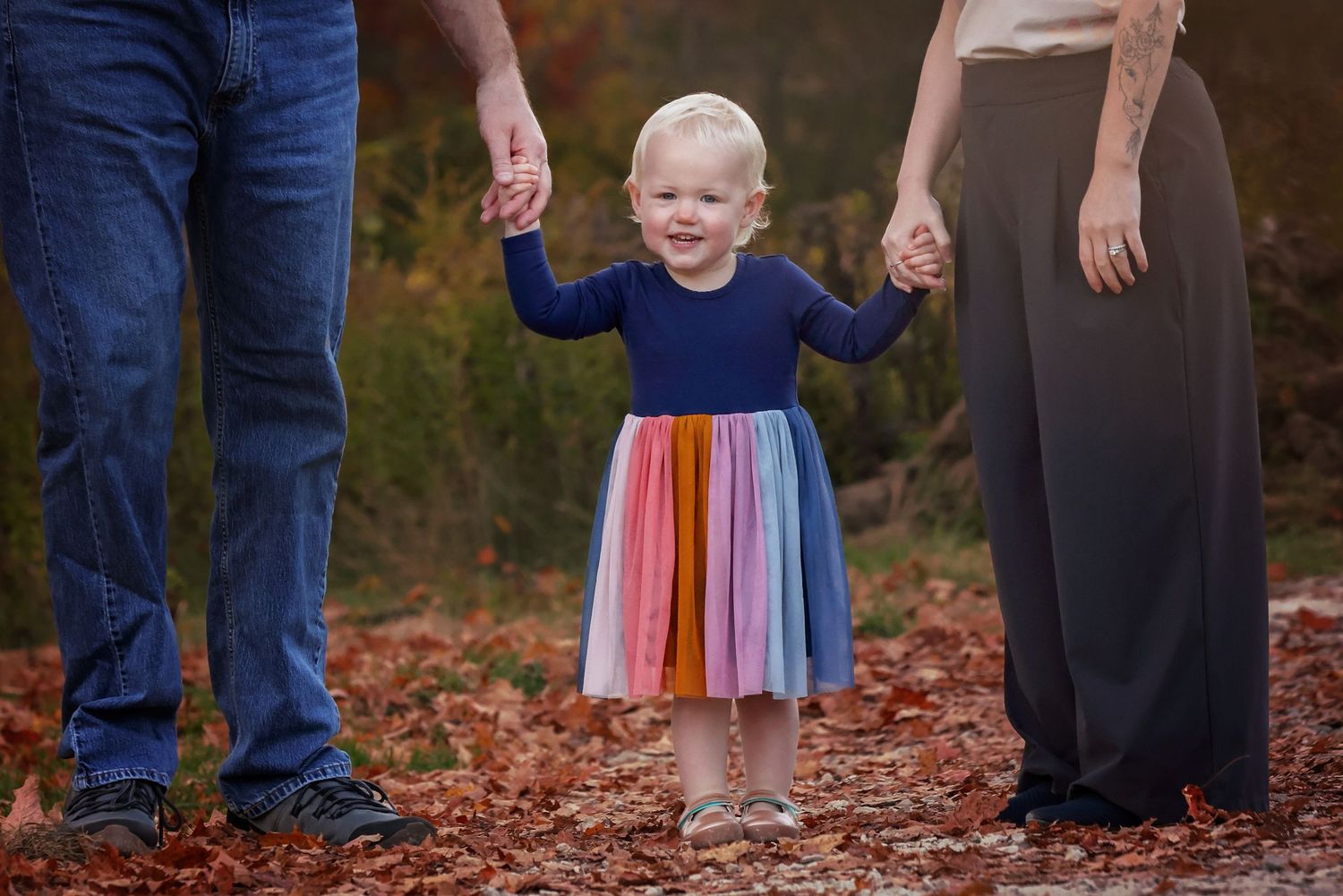 A small child in a colorful rainbow tulle skirt stands among fallen autumn leaves in a wooded setting.