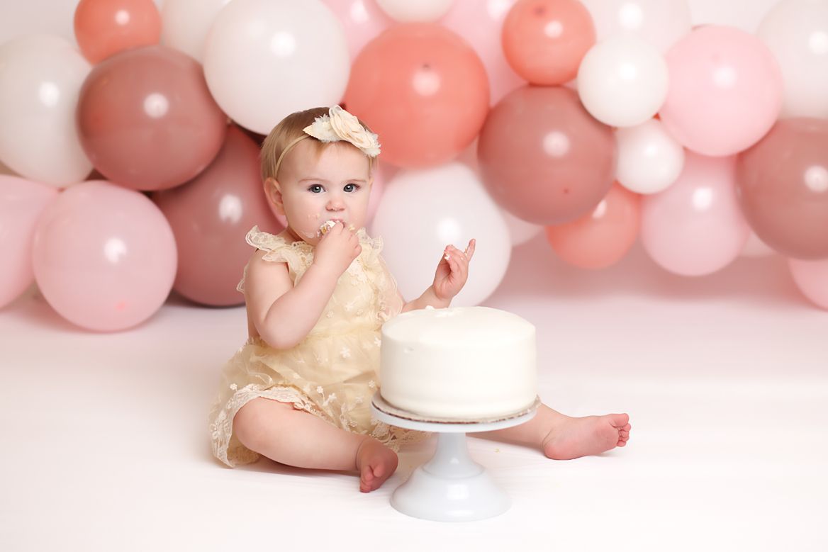 Baby poses in white bathtub with pink and peach balloons for cake smash photography session.