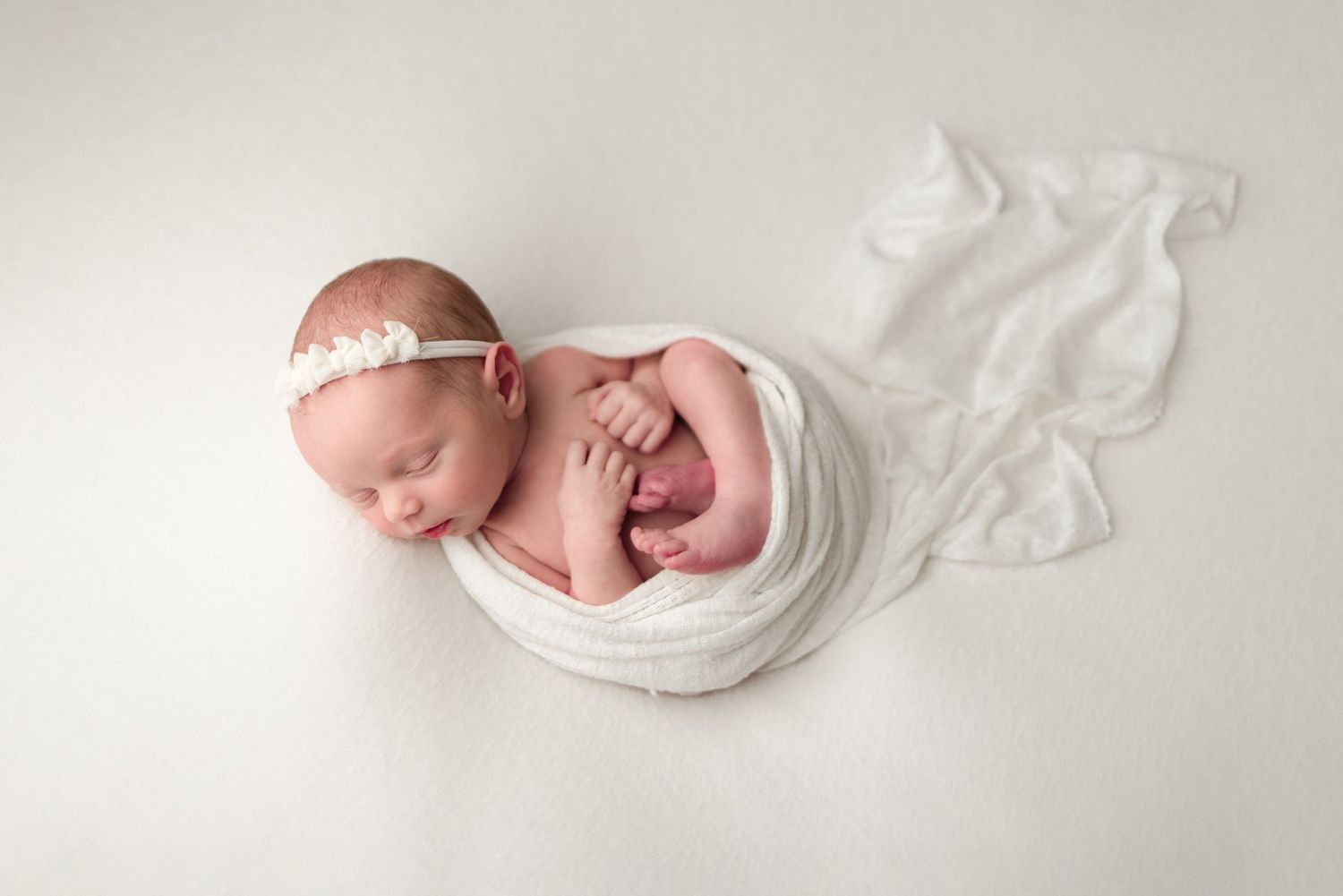 A sleeping newborn baby swaddled in white fabric lies peacefully against a light backdrop.