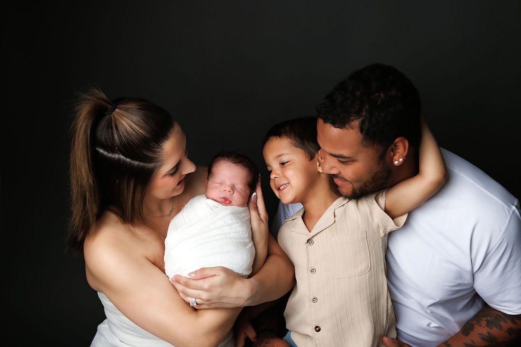 Family of four posing for newborn portrait, parents and toddler smiling at swaddled baby against dark background.