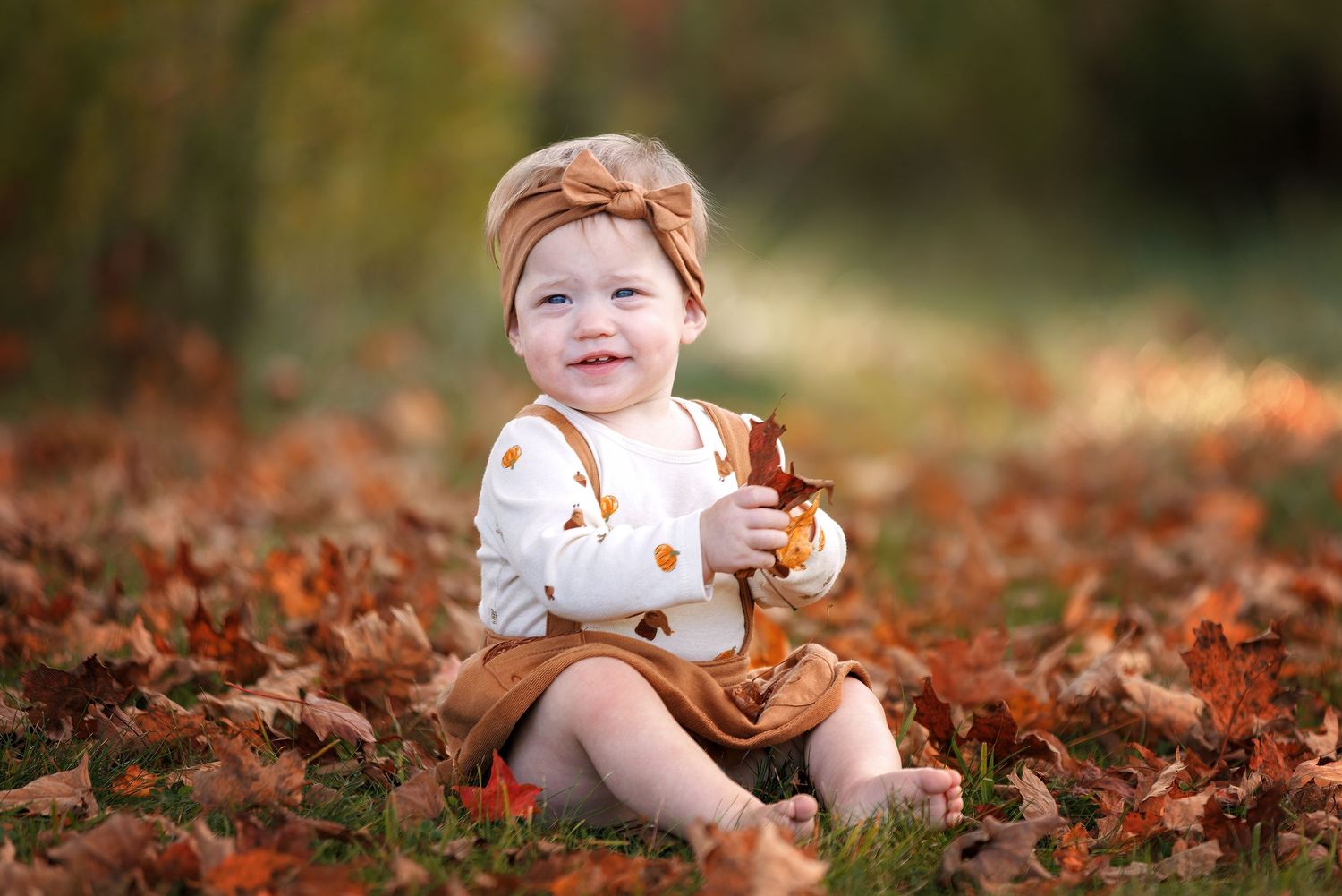 A child in autumn attire sits among vibrant orange and red fallen leaves while holding a small pumpkin.