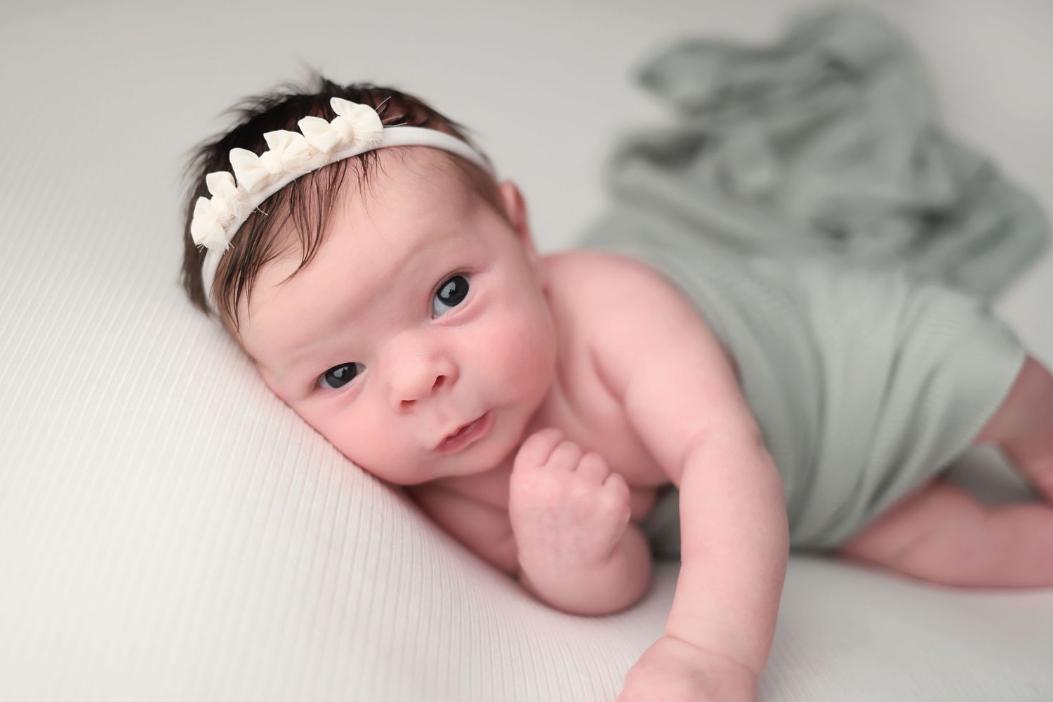 Baby wearing mint green clothing lies on white bedding while making various sweet expressions.