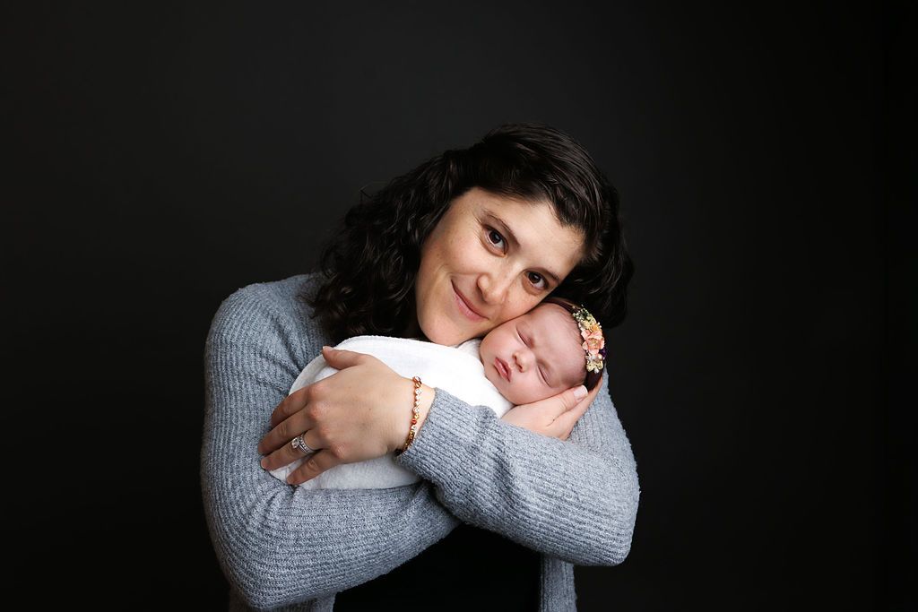Mother tenderly holds sleeping swaddled newborn baby close to her chest on dark background.