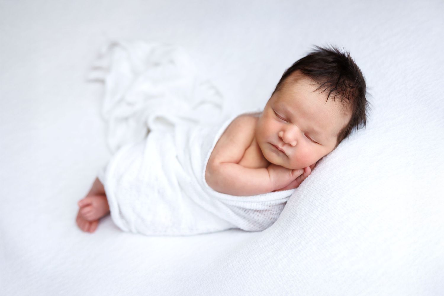 Peaceful newborn baby swaddled in white wrap sleeping on soft white blanket in professional photo.