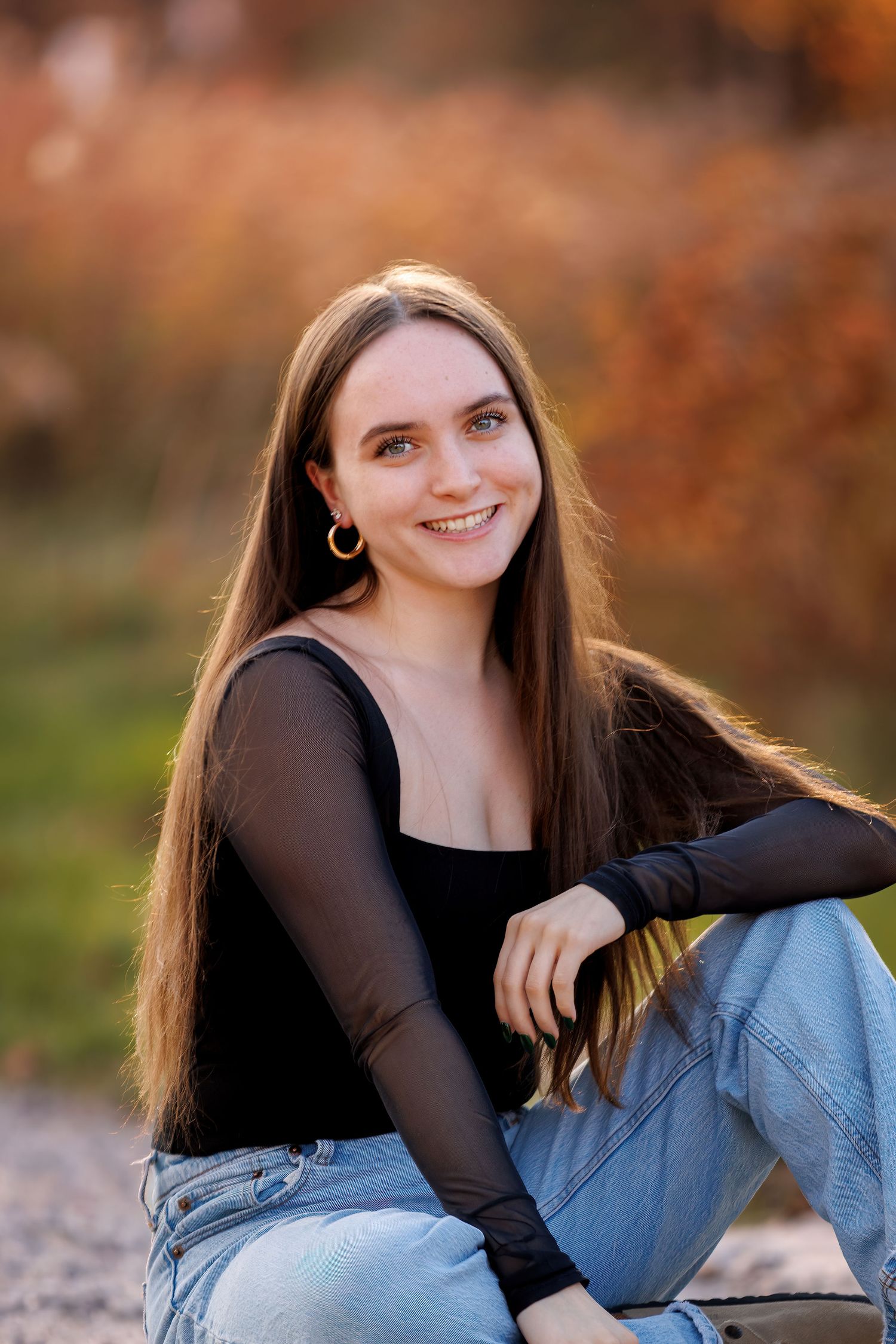 Young woman with long brown hair smiling outdoors, wearing black top and jeans, seated against autumn foliage background.