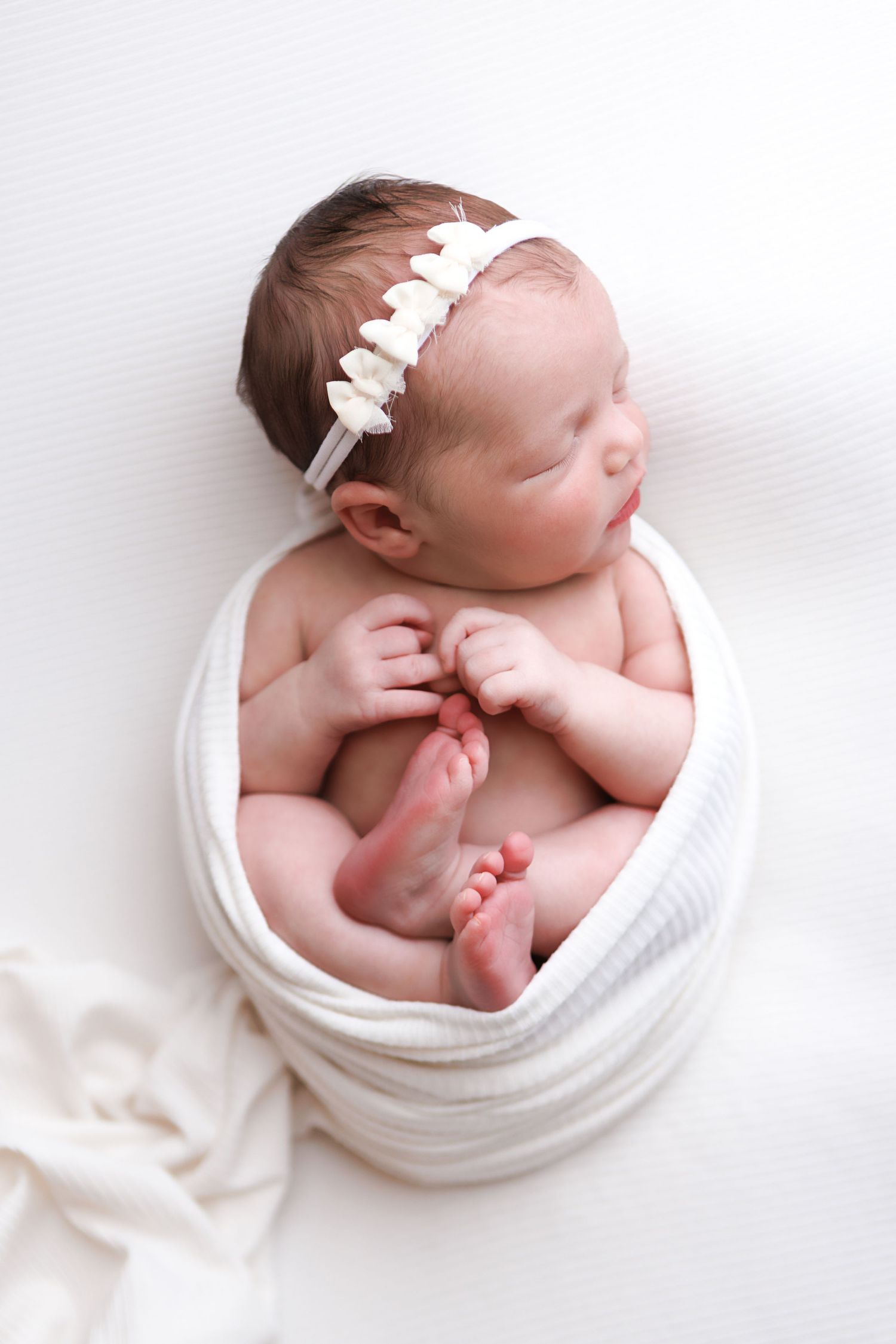 Sleeping newborn baby girl wrapped in white swaddle blanket wearing a delicate floral headband on white background. Windham NH Newborn.