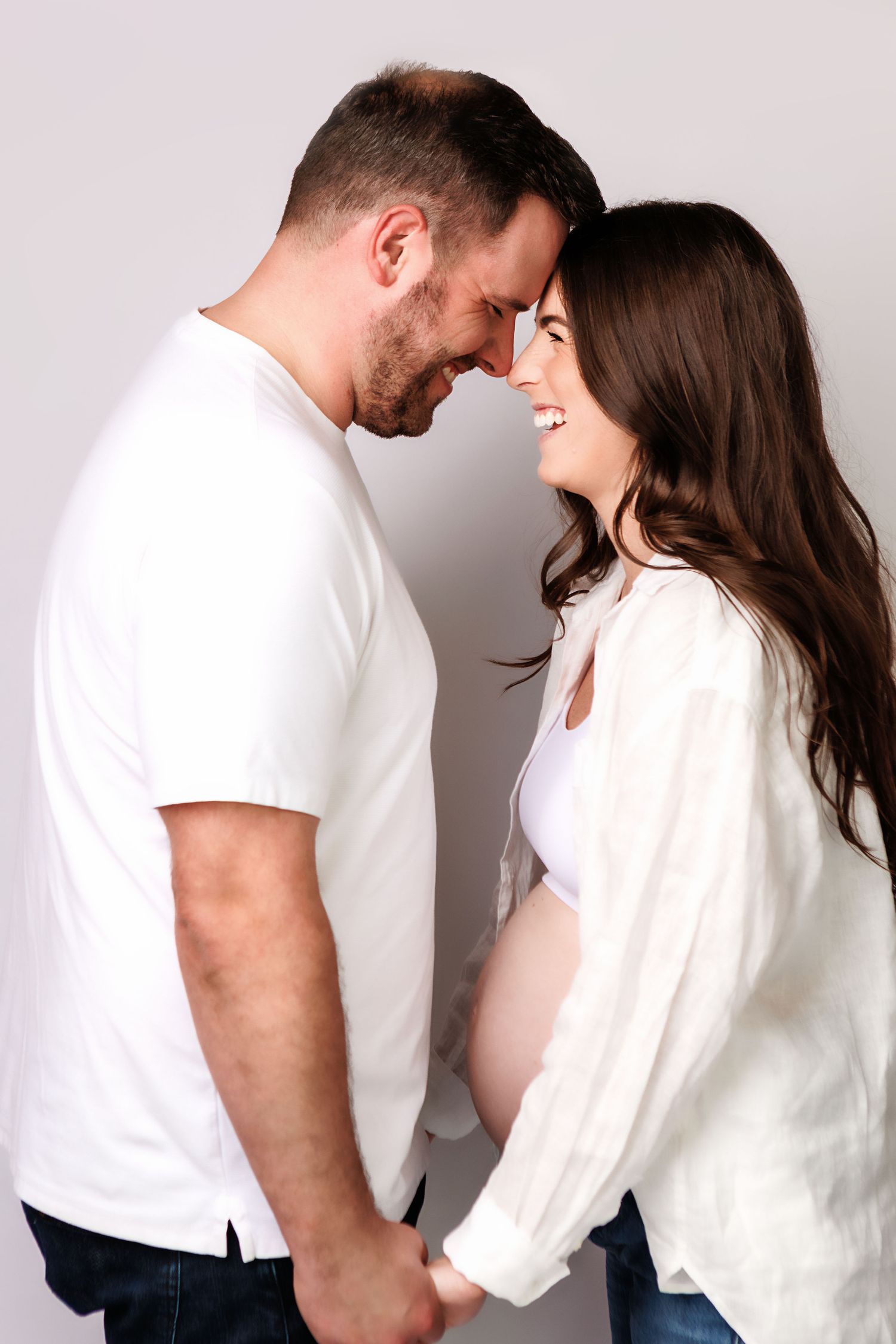 Happy pregnant couple holding hands and touching foreheads, smiling together against a white background.