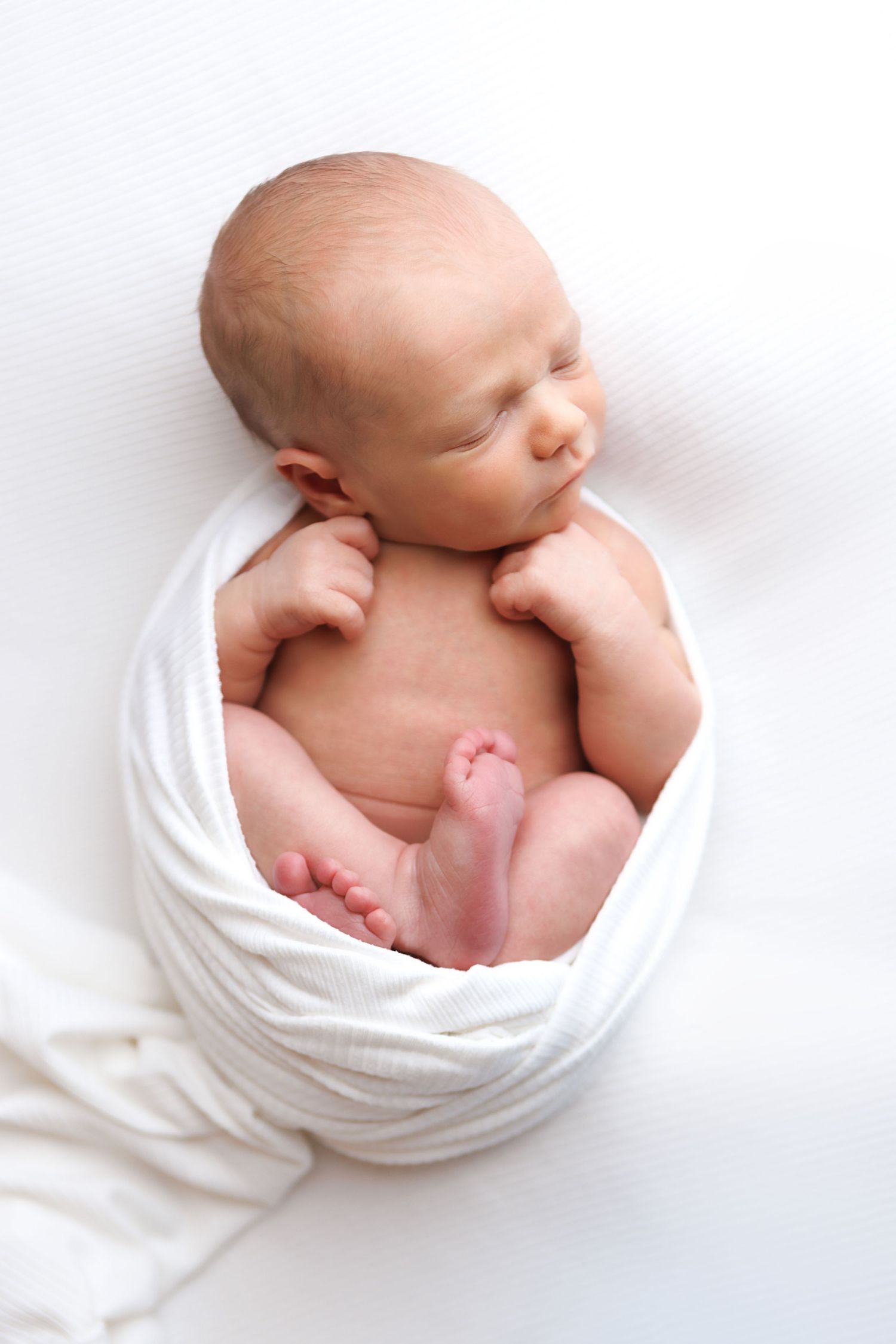 Peaceful newborn baby swaddled in white wrap, sleeping curled up on a soft white background.