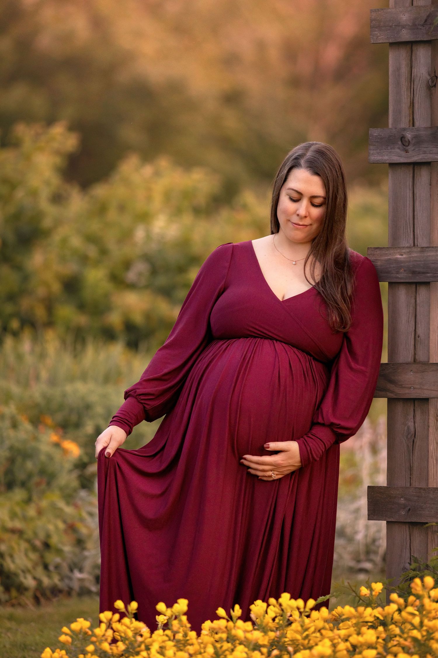 A woman in a burgundy dress stands among yellow wildflowers during a maternity photoshoot at sunset.