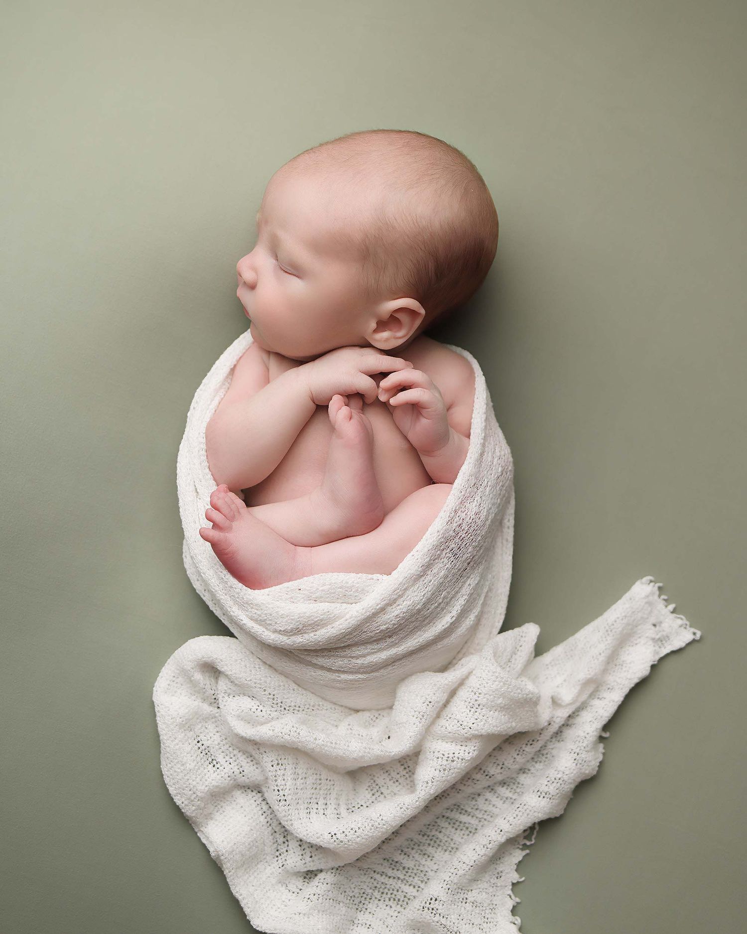 Artistic newborn portrait series with baby wrapped in white textured blanket against sage background.