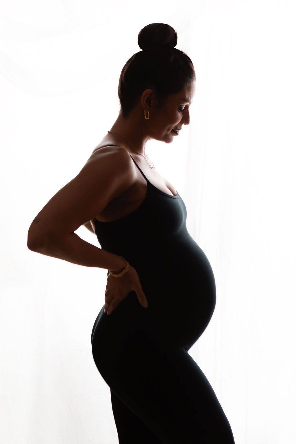Side profile silhouette of pregnant woman in black dress holding lower back by window.
