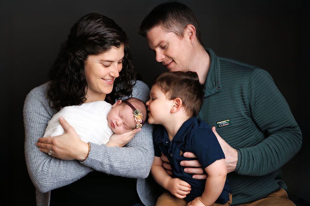 Family portrait with parents and toddler son kissing their sleeping newborn baby girl in a studio.