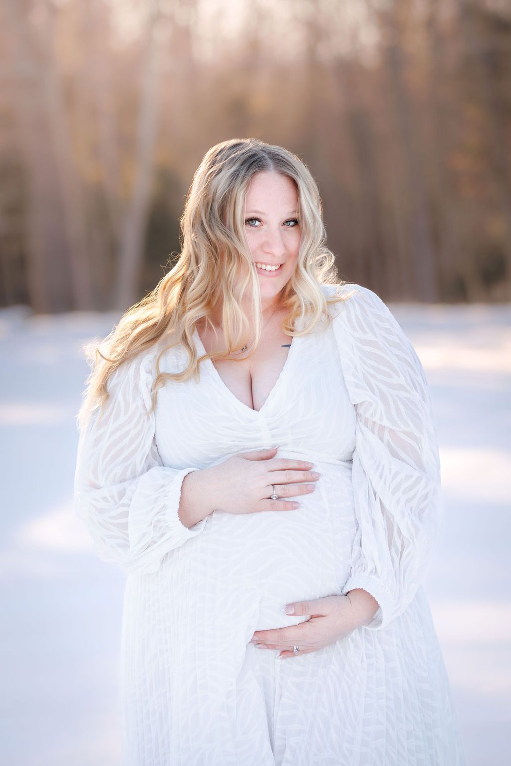 Smiling pregnant woman in white lace dress holding baby bump outdoors in snowy winter landscape.